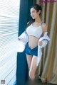 A woman in a white top and blue shorts posing by a window.