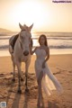 A woman in a white dress standing next to a white horse on the beach.